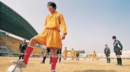 Film still from Shaolin Soccer (2001). Image of a monk stopped, his foot on a soccer ball, preparing to make a goal.