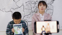 Film still from Total Trust (2023). Image of a woman and son being filmed on a phone, the boy holding an iPad with an image of his father.